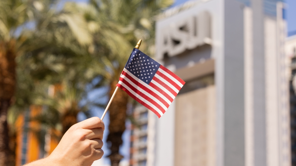 A hand holds a tiny American flag in front of an ASU®168极速入口一分钟赛车网 sign