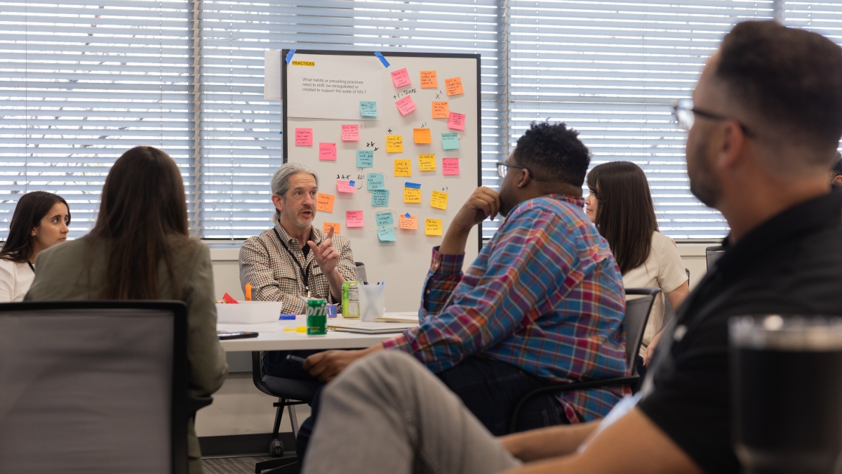 A seated man speaks to a room of people about his ideas with a whiteboard of sticky note ideas behind him.
