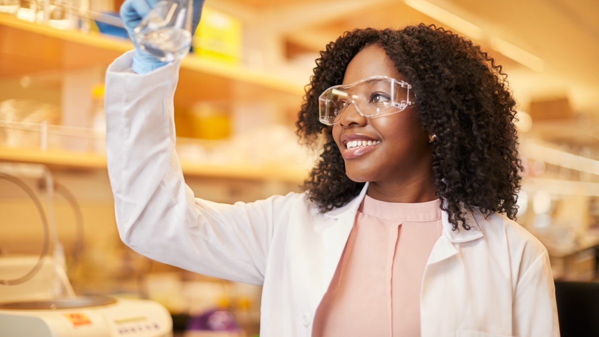 Young Black woman wearing lab coat and glasses looks at a beaker